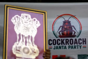The Cockroach Janta Party (CJP) logo is pictured in the background as a supporter holds India's national emblem during a protest over alleged irregularities in the country's major examinations, in New Delhi. - AFP